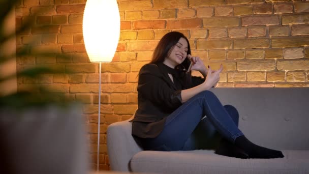 Retrato de la joven morena caucásica sentada en el sofá y hablando en el teléfono celular alegremente en el ambiente del hogar . — Vídeos de Stock