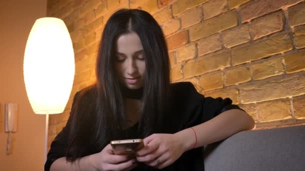 Портрет юной брюнетки кавказской девушки, сидящей на диване и смотрящей в смартфон улыбается в камеру в домашней обстановке . — стоковое видео