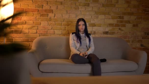 Nahaufnahme einer jungen hübschen kaukasischen Frau, die im Fernsehen lächelt und fröhlich lacht, während sie drinnen auf der Couch sitzt — Stockvideo