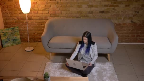 Κοντινό πλάνο του νεαρού όμορφο καυκάσιο θηλυκό βλέποντας μια ταινία στο φορητό υπολογιστή που κάθεται στο πάτωμα σε ένα φιλόξενο διαμέρισμα με μοντέρνους εσωτερικούς χώρους — Αρχείο Βίντεο