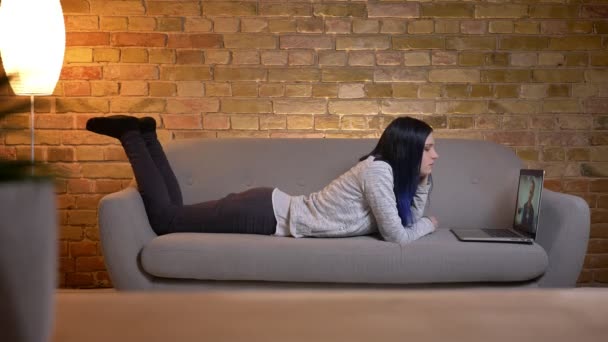 Närbild porträtt av unga vackra kaukasiska hona med färgat hår tittar på en film på den bärbara datorn medan liggande på soffan inomhus — Stockvideo