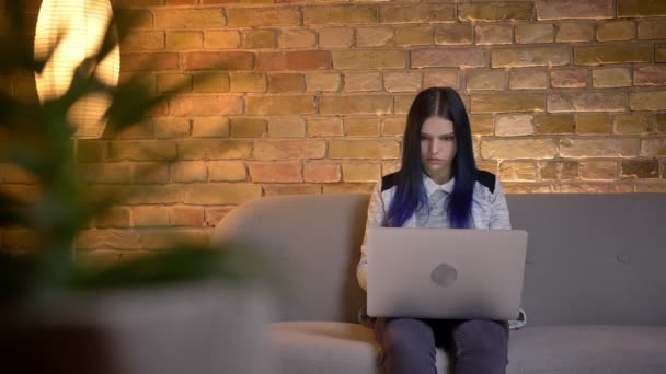 Κοντινά πλάνα της νεαρής όμορφη Καυκάσιος θηλυκό μηνύματα στο φορητό υπολογιστή, ενώ κάθεται στον καναπέ στο άνετο σπίτι σε εσωτερικούς χώρους — Αρχείο Βίντεο