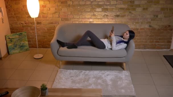 아늑한 아파트에서 소파에 누워 있는 동안 태블릿을 사용 하 여 젊은 예쁜 백인 소식통 여성의 근접 촬영 — 비디오