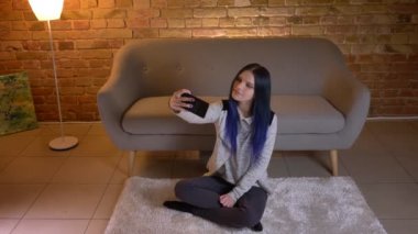 Genç çekici beyaz kadın seyir portre telefonda selfie 'ler alarak ve rahat evde kapalı yerde otururken gülümseyen