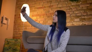 Genç güzel beyaz hipster kadın çekim çekim telefonda selfie 'ler yapmak ve rahat evde kapalı yerde otururken kameranın önünde gülümsüyor