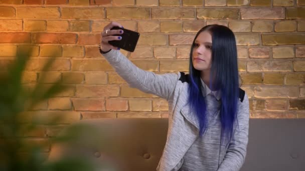 Zbliżenie strzelać z młody ładny kaukaski Hipster żeński z farbowane włosy biorąc selfie na telefon siedzi na kanapie w przytulnym domu wewnątrz — Wideo stockowe