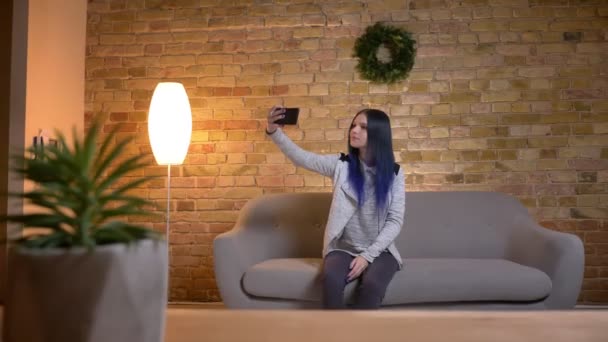 Крупним планом знімок молодої привабливої кавказької хіпстерки, яка приймає селфі на телефон, сидячи на дивані в затишній квартирі в приміщенні — стокове відео