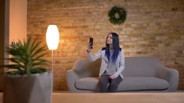 Closeup shoot of young attractive caucasian hipster female taking selfies on the phone and smiling while sitting on the couch at cozy home indoors — Stock Video