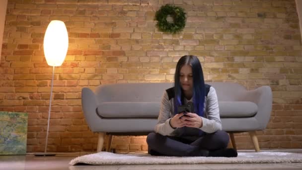 Zbliżenie strzelać z młody ładny kaukaski Hipster żeński z farbowane włosy wpisując na telefon siedząc na podłodze w przytulnym mieszkaniu w pomieszczeniu — Wideo stockowe
