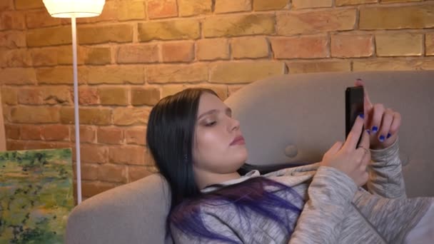 Primer plano de la joven hipster caucásica mujer navegando por la web en el teléfono tumbado en el sofá en un acogedor apartamento en el interior — Vídeo de stock