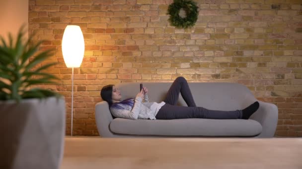 Съемки крупным планом молодой привлекательной кавказской хипстерки с телефона, лежащей на диване в уютном доме — стоковое видео
