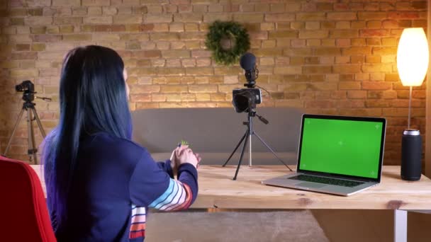 Съемки крупным планом молодой привлекательной кавказской блогерки, транслирующей разговор на камеру и имеющей перед собой ноутбук с зеленым экраном — стоковое видео