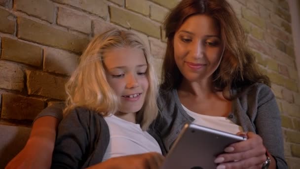 Close-up opnamen van de jonge Kaukasische moeder en haar kleine mooi meisje met behulp van de tablet samen zittend op de Bank thuis binnenshuis — Stockvideo