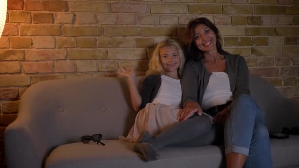 Closeup πυροβολούν νεαρή μητέρα και η κόρη της βλέποντας μια κωμωδία στην τηλεόραση μαζί να κάθονται σε καναπέ αγκάλιασμα και χαμογελαστός — Αρχείο Βίντεο