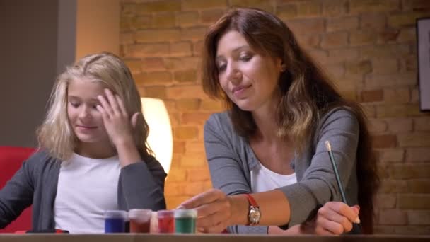 Съемки крупным планом молодой женщины и ее маленькой красавицы-дочери, рисующей вместе и осматривающей краски — стоковое видео