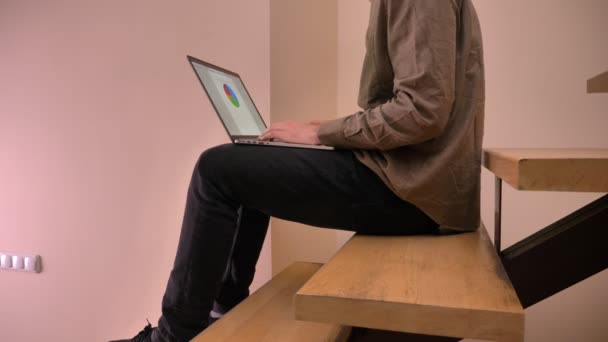 Close-up van de mens te typen op de laptop met diagram op het scherm zitten in het kantoor binnen schieten — Stockvideo