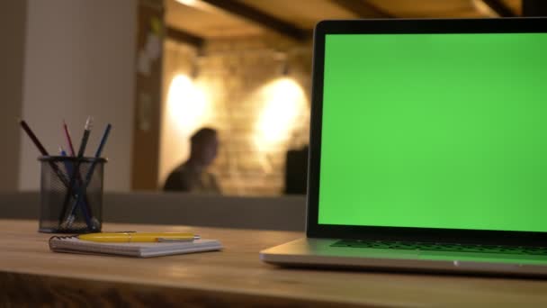 Primer plano de la pantalla verde de la computadora portátil acostado en el escritorio junto a la taza con lápices en el interior de la oficina con un empleado en el fondo — Vídeos de Stock