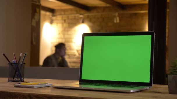 Närbild skott av en grön chroma skärm på laptop liggande på skrivbordet på kontoret inomhus med en anställd arbetar på den suddig bakgrunden — Stockvideo