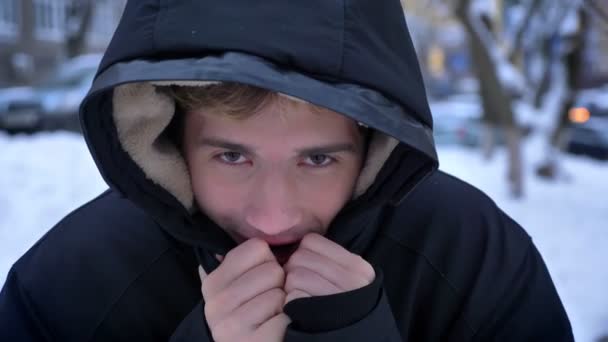 Primer plano del joven guapo caucásico congelándose y calentándose sonriendo en un abrigo de invierno al aire libre en un día nevado — Vídeos de Stock