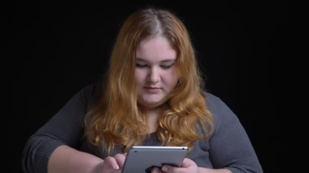 Съемки крупным планом молодой белой женщины с избыточным весом, печатающей на планшете, затем смотрящей в камеру и улыбающейся — стоковое видео