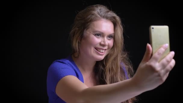 Närbild porträtt av mellersta åldern kaukasiska brunett kvinna med ett videosamtal om telefonen viftande hello med en hand och ler glatt — Stockvideo