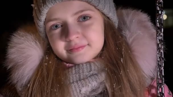 Retrato de cerca de la pequeña chica bonita montando lentamente en el columpio y mirando con gusto en la cámara en el fondo de la calle de invierno . — Vídeo de stock