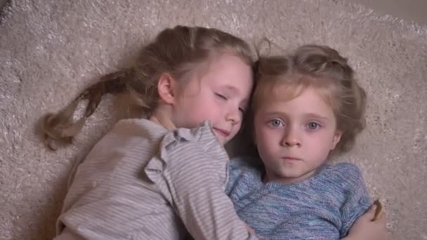 Top shot portrait of two small funny girls hugging and kissing joyfully with each other and lying on the floor. — Stock Video