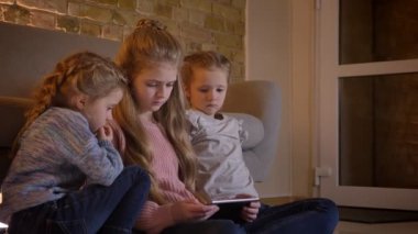 Portre profil, katta oturan ve içine tablet rahat ev atmosferde dikkatle izliyor üç oldukça beyaz kız.