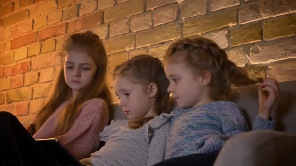 Portrait de trois jolies filles caucasiennes assises sur le canapé et regardant attentivement dans la tablette dans une atmosphère confortable à la maison . — Video