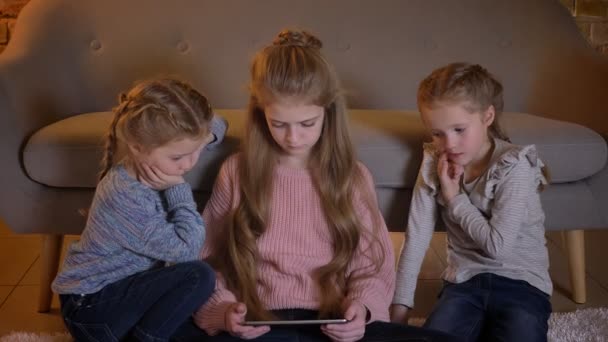 Portrait de trois jolies filles caucasiennes assises sur le sol et regardant attentivement dans la tablette dans une atmosphère confortable à la maison . — Video