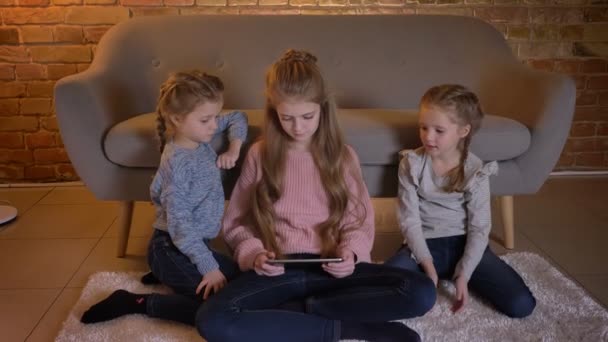 Porträtt av tre ganska vita tjejer sitter på golvet och titta in i tablett på allvar i mysiga hem atmosfär. — Stockvideo