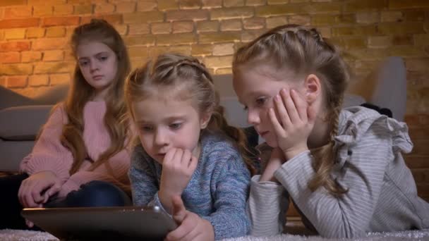 Portret van drie kleine Kaukasische meisjes op de vloer liggen en kijken naar tablet aandachtig in gezellige thuis. — Stockvideo