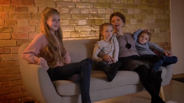 Retrato familiar de madre caucásica con tres hijas sentadas juntas y viendo la película riéndose en un acogedor ambiente hogareño . — Vídeo de stock