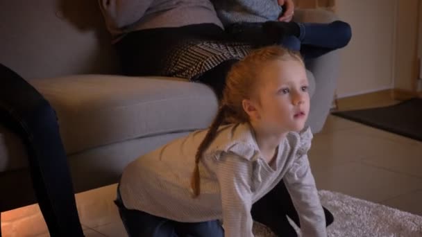 Close-up retrato de pequena menina caucasiana com tranças assistindo filme atentamente e apontando o dedo sobre ele com sua mãe e irmã no fundo . — Vídeo de Stock