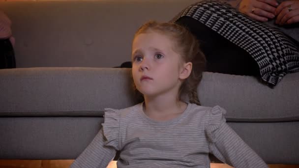 Nahaufnahme Porträt eines kleinen kaukasischen Mädchens mit Zöpfen beim aufmerksamen Kinobesuch in gemütlicher Atmosphäre zu Hause. — Stockvideo