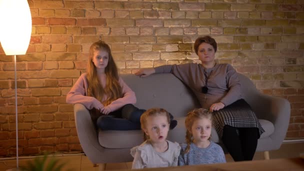 Family portrait of caucasian mother with three daughters sitting on sofa and watching movie in cosy home atmosphere. — Stock Video