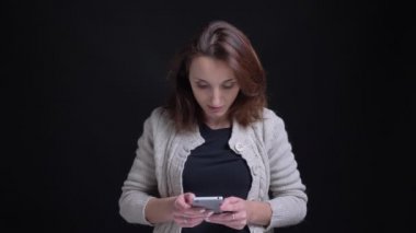 Orta yaşlı esmer beyaz kadın gülümseyerek siyah arka plan üzerine smartphone içine izlerken portresi.