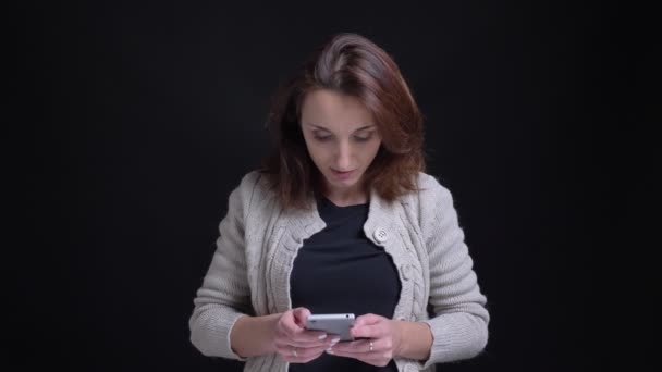 Portrait of middle-aged brunette caucasian woman watching into smartphone smilingly on black background.