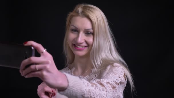 特写镜头拍摄的中年白种人金发女郎有一个视频通话在电话里微笑着, 愉快地说话与背景隔离在黑色 — 图库视频影像