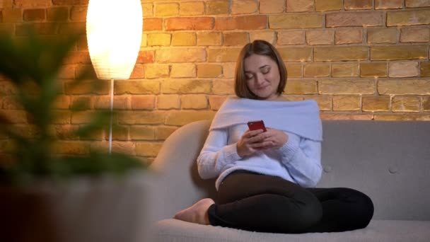Крупный план съемки молодой веселой кавказской женщины, переписывающейся по телефону и счастливо улыбающейся, расслабляющейся дома — стоковое видео