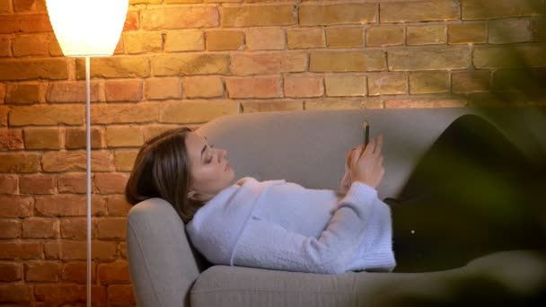 Closeup portret van de jonge mooie Kaukasische vrouwelijke liggend op de Bank en SMS op de telefoon binnenshuis thuis gezellig — Stockvideo