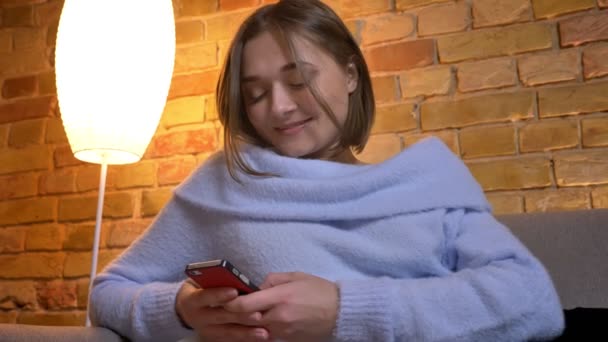 Closeup shoot of young cute caucasian female texting a message on the phone fixing her hair and smiling at cozy home indoors — Stock Video