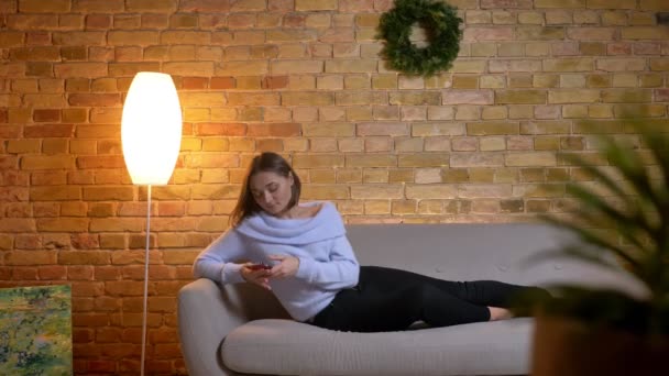 Крупним планом портрет молодої короткошерстої жінки, використовуючи телефон, відпочиваючи на дивані в приміщенні — стокове відео