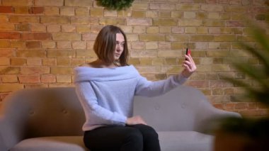 Closeup çekim genç sevimli beyaz kadın poz ve saçı kapalı rahat evde kanepede otururken sabitleme telefonda selfies alarak