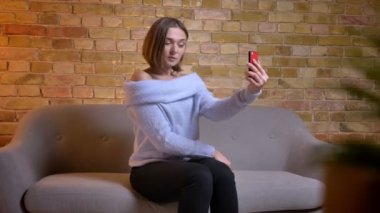Closeup çekim genç sevimli beyaz kadın kapalı rahat evde kanepede otururken telefonda selfies alarak