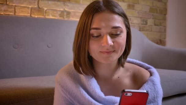 Closeup střílet z mladé okouzlující Kavkazský žena pomocí telefonu při pohledu na fotoaparát a usmívá se na útulný domov uvnitř — Stock video