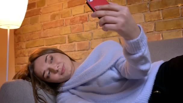 Closeup shoot of young beautiful caucasian female lying on her side on the couch and taking selfies on the phone being relaxed at home — Stock Video