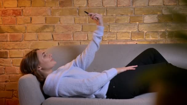 Primer plano de brote de joven caucásica hembra tomando selfies en el teléfono mientras está acostado en el sofá en el acogedor hogar en el interior — Vídeo de stock