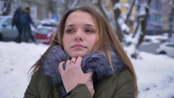 Closeup portret van jonge aantrekkelijke Kaukasische vrouwtje met donkerbruine haren bevriezen en verstopt in een gezellige winterjas in een besneeuwde dag — Stockvideo