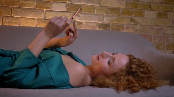 Närbild porträtt av ingefära curly-haired flicka liggande på soffan och titta på i din smartphone på mysiga hem bakgrund. — Stockvideo